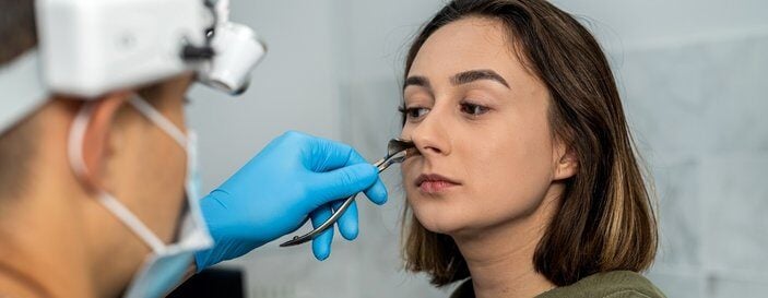 ENT specialist wearing a headlight and blue gloves examining a young woman's nose with a medical instrument in a clinic