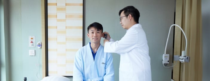 身穿白袍的医生在现代化医院病房内使用耳镜检查男性病人的耳朵。病人身穿浅蓝色浴袍,坐在病床上微笑。 身穿白袍的医生在现代化医院病房内使用耳镜检查男性病人的耳朵。病人身穿浅蓝色浴袍,坐在病床上微笑。