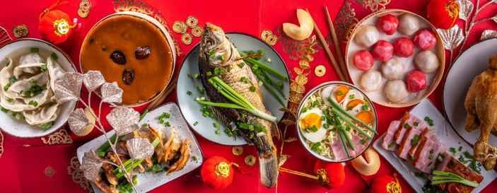 Festive Chinese New Year feast with traditional dishes, including whole steamed fish, dumplings, tangyuan, braised pork, and other symbolic foods on a red tablecloth decorated with gold coins and lanterns.