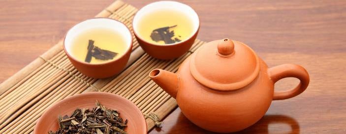 Traditional Chinese tea ceremony setup with a clay teapot, two teacups filled with brewed tea, and a plate of loose tea leaves on a bamboo mat.