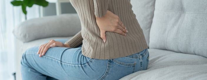 a woman sitting on a grey couch, holding her lower back in pain, possibly experiencing backache or kidney discomfort.