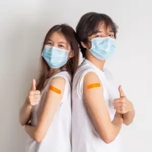 Two women wearing face masks showing vaccination bandages on their arms.